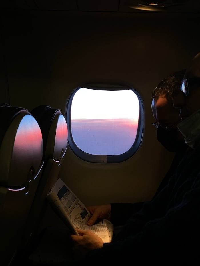 A passenger reads a magazine beside a sunset view from the airplane window, capturing a serene travel moment.